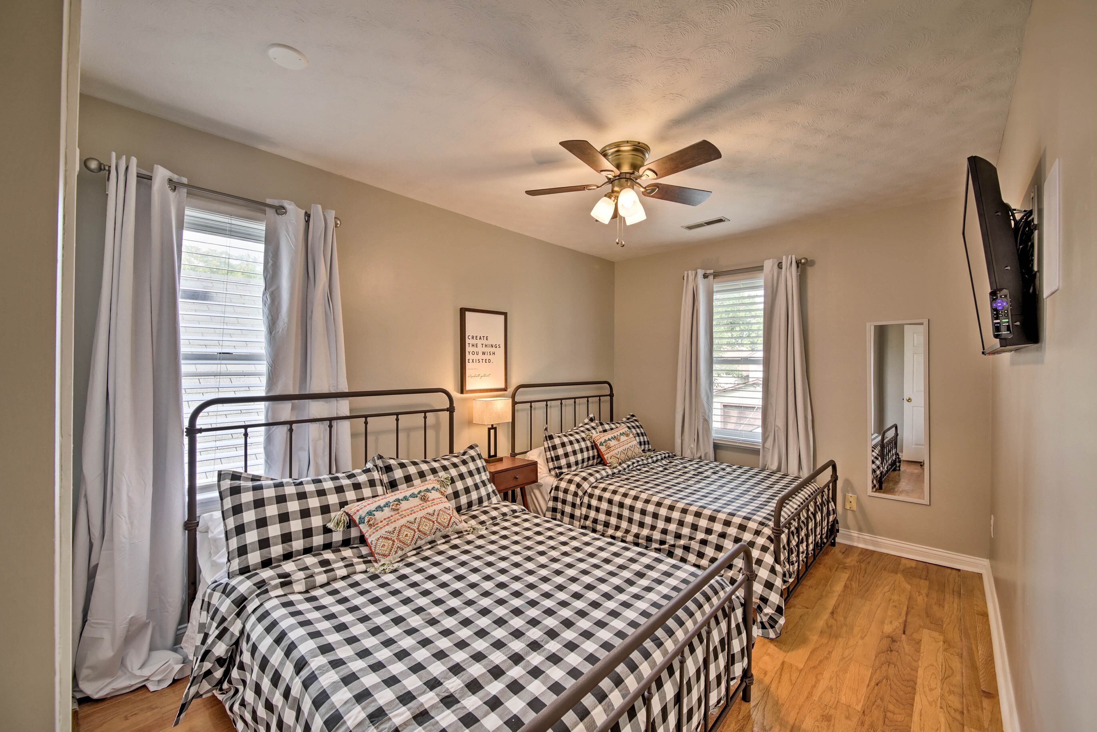 A bedroom with two beds, a ceiling fan, a bedside table with a lamp, and a wall-mounted TV.
