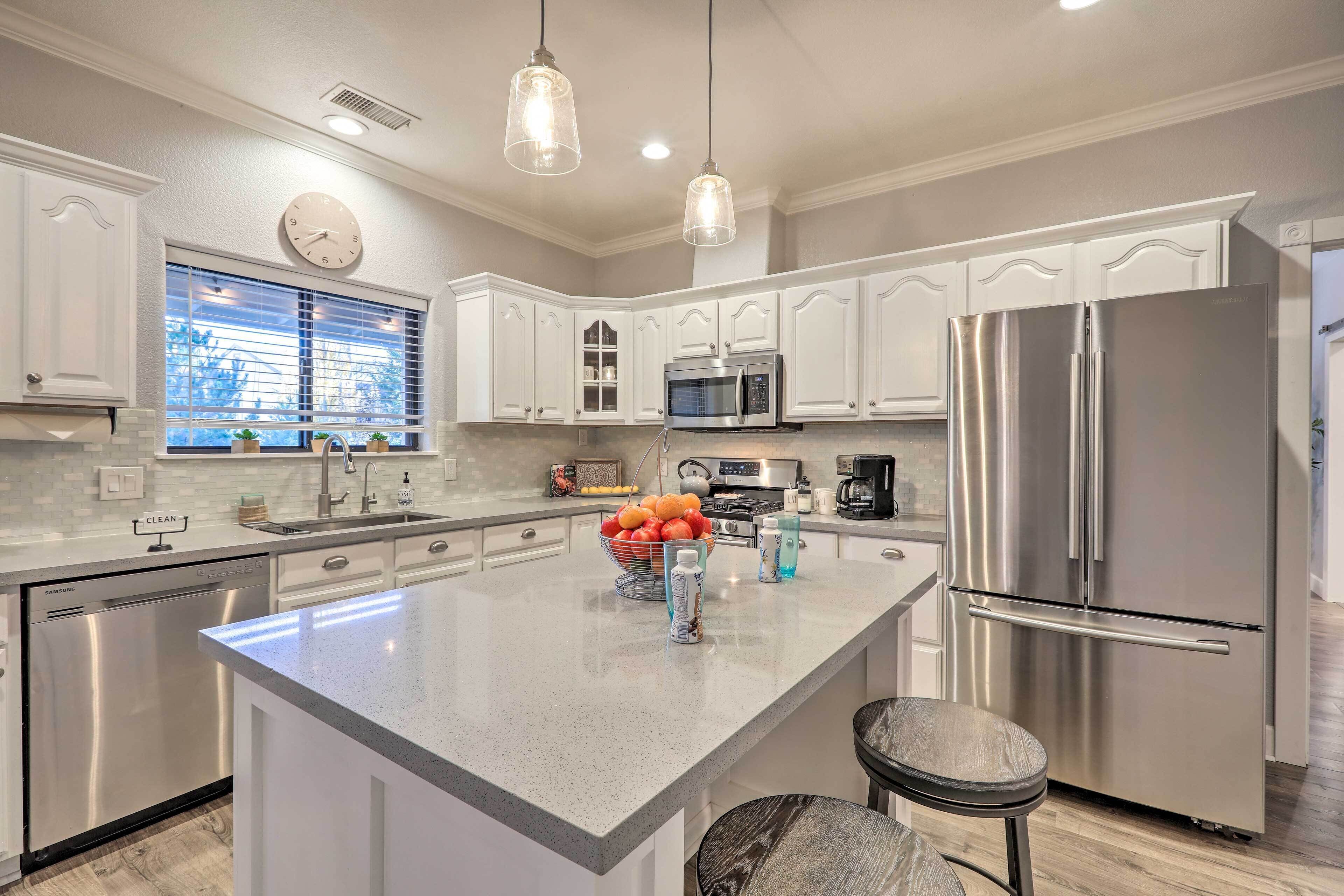 A modern kitchen with white cabinetry, stainless steel appliances, a central island with fruit, and pendant lighting.