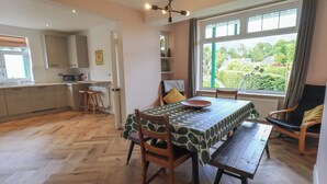 A dining area with a herringbone wooden floor, a table with a leaf-patterned tablecloth, and a view of a garden through a large window. - Limhus House (Keswick)