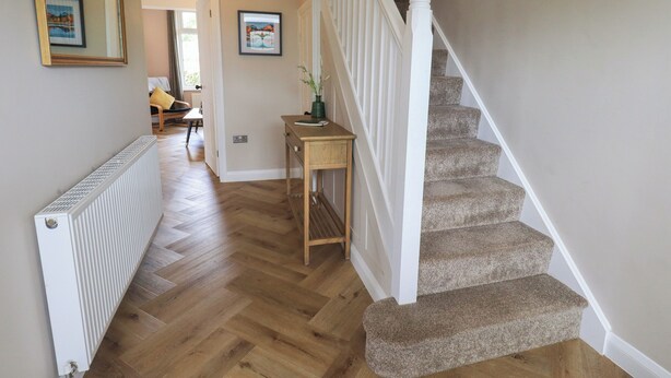 A hallway with a wooden floor, a white radiator, a small table with a plant, and a staircase with carpeted steps. - Limhus House (Keswick)