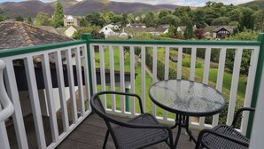 A balcony with a glass table and two chairs, overlooking a green landscape and houses. - Limhus House (Keswick)