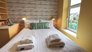 A neatly made bed with two folded towels, a wooden wall, a window with a view, and a small desk with a lamp. - Limhus House (Keswick)
