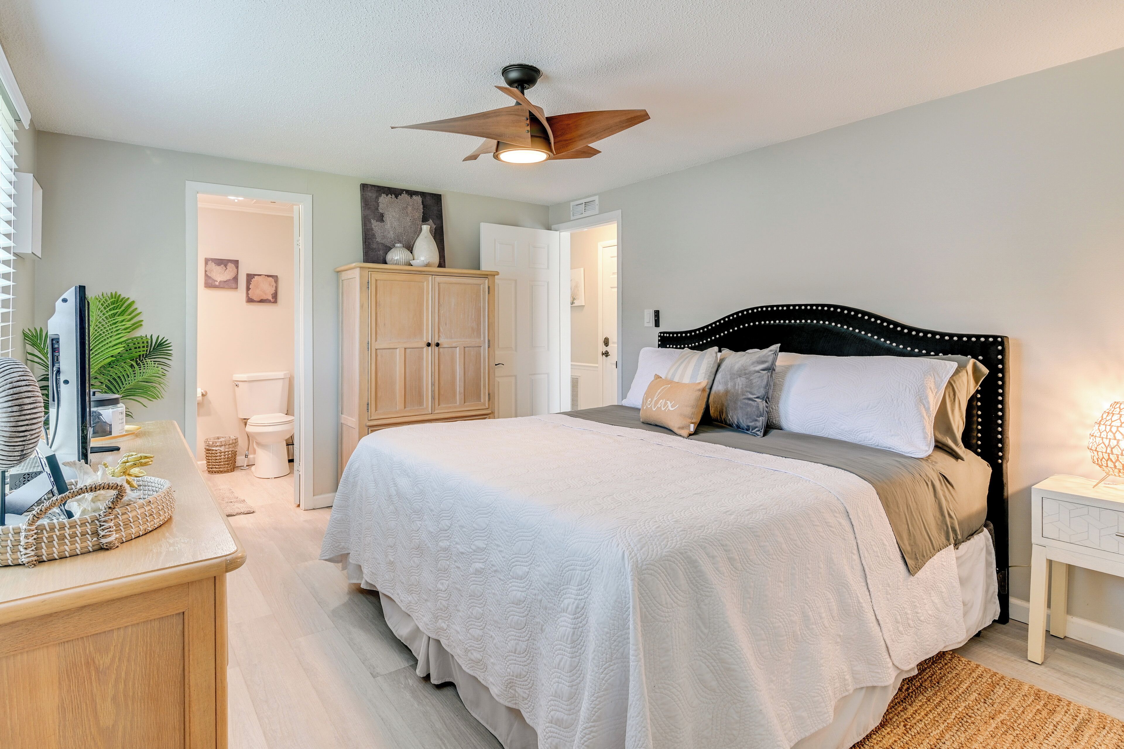 A bedroom with a bed, bedside table, nightstand, ceiling fan, and a door leading to another room.
