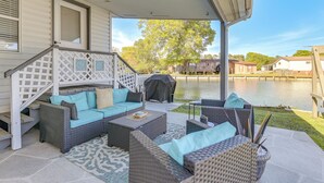 A covered outdoor patio with wicker furniture, a covered grill, and a view of a lake and houses in the background.