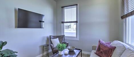 A living room with a sofa, a coffee table, a chair, a TV mounted on the wall, and a window with blinds.