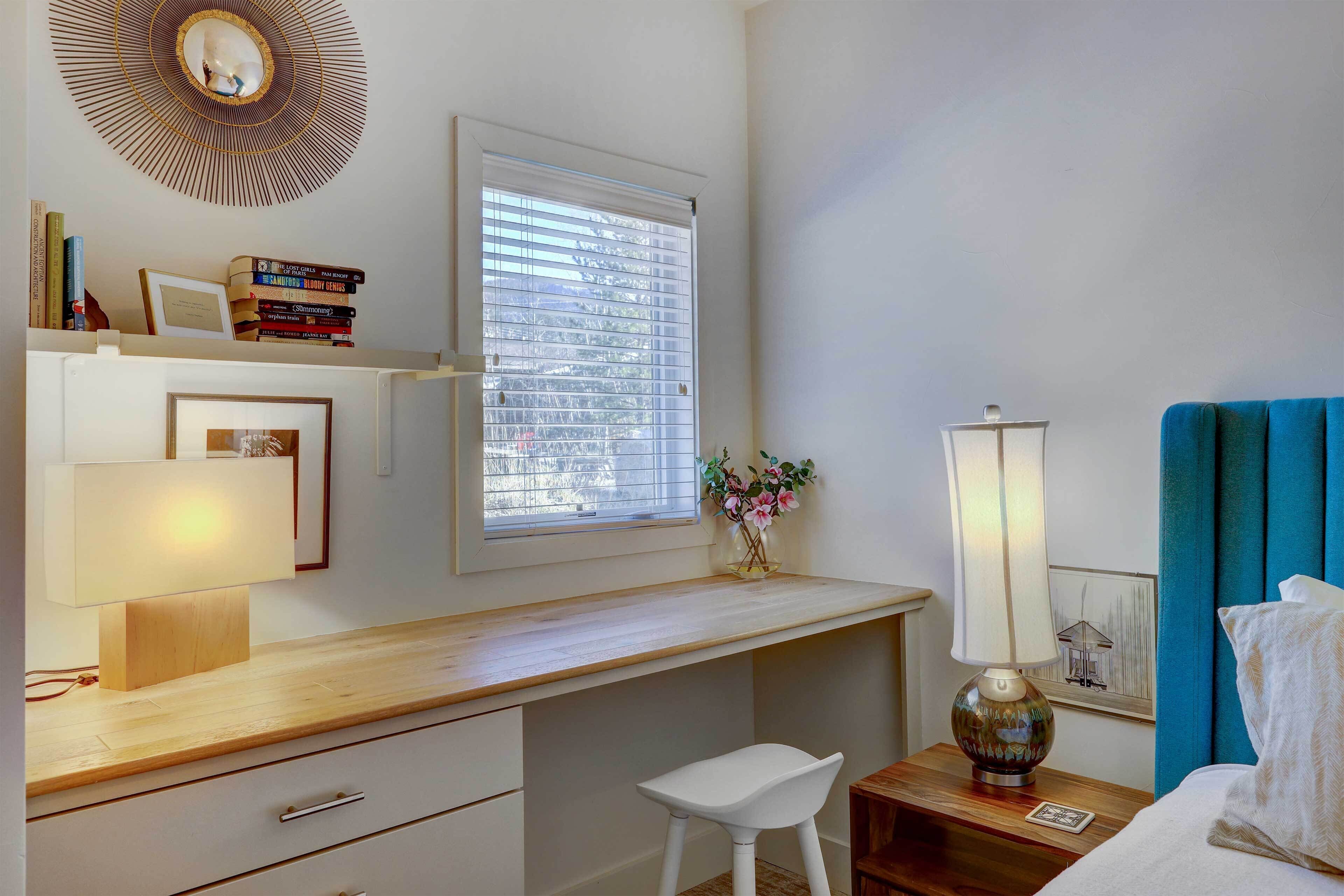 Une chambre avec un bureau en bois, une chaise blanche, une table de chevet avec une lampe, un lit avec une tête de lit bleue et une fenêtre avec des stores.
