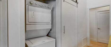 A laundry room with a front-loading washer and dryer, a sliding door closet, and a wooden floor.