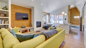 A modern living room with a yellow sofa, wooden panel walls, a flat-screen TV, and a dining area with a kitchen in the background.