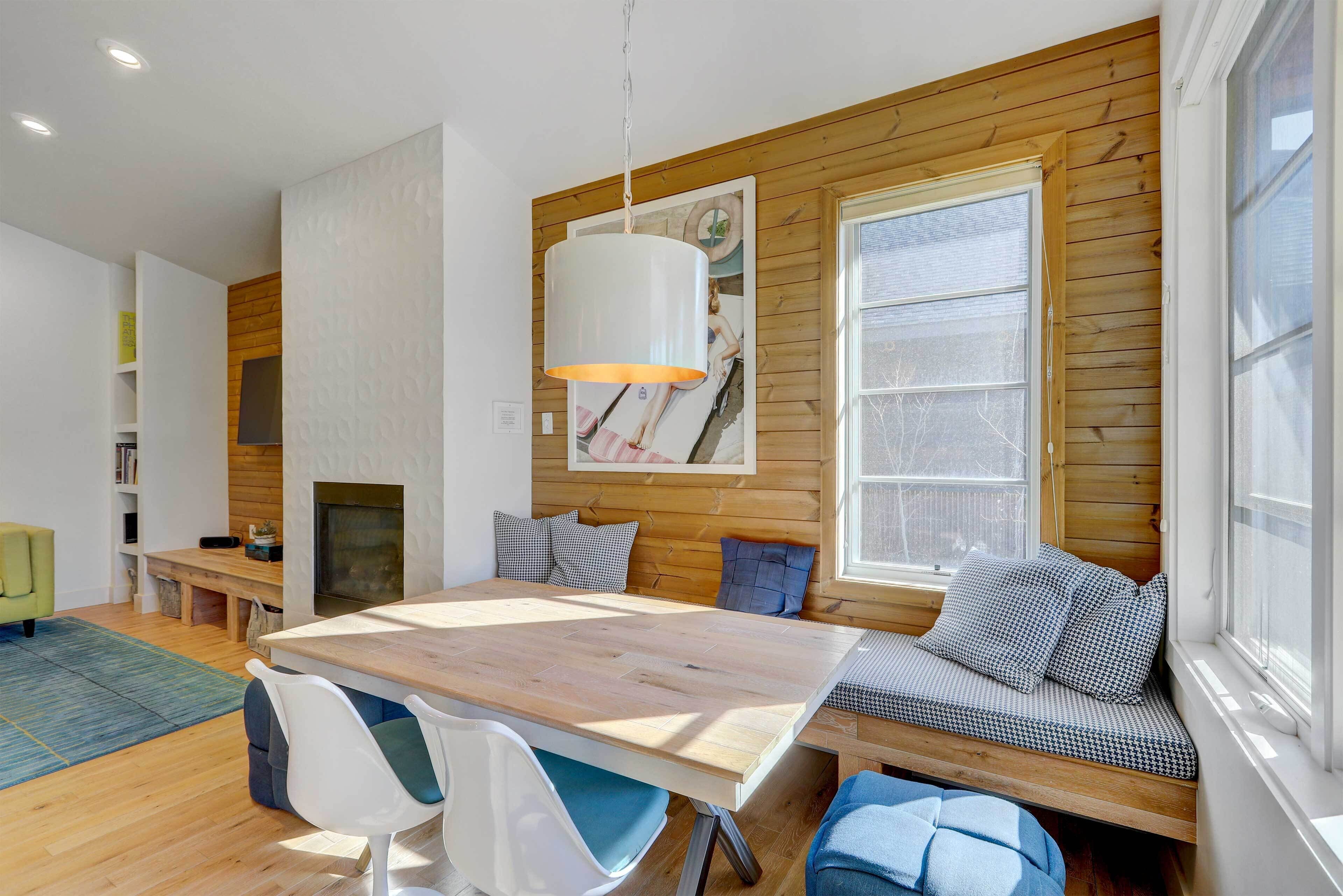 A modern living room with a wooden wall, a large window, a dining table, and a built-in bench with cushions.