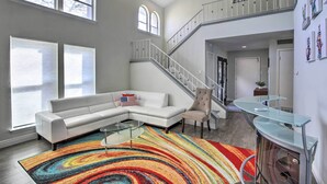 A spacious living room with a white sectional sofa, a colorful rug, and a glass coffee table. A staircase with a glass railing leads to an upper level. - Movie Room & Private Pool: Richardson Retreat (Richardson)