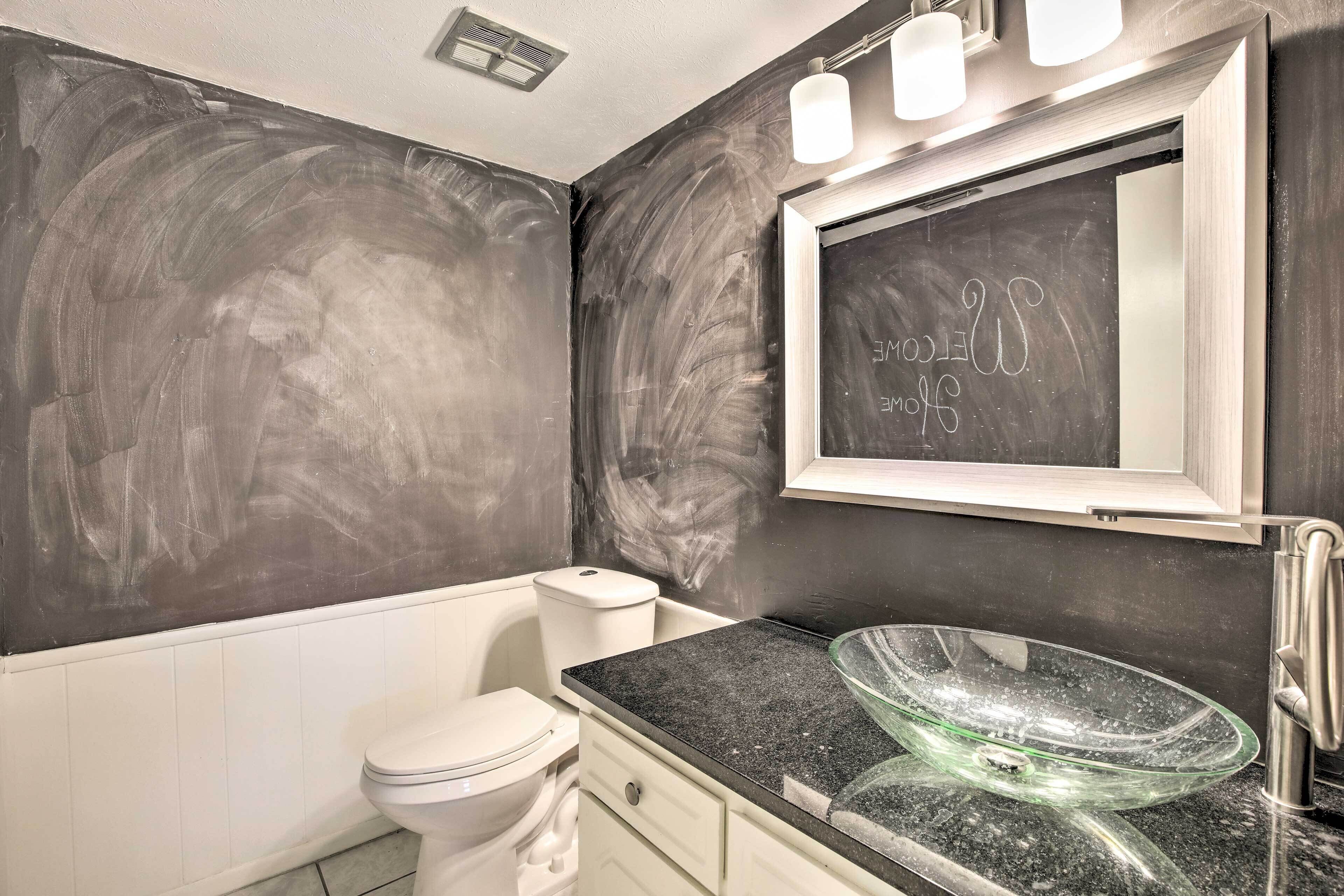 A bathroom with a black and white tiled wall, a white toilet, a glass bowl sink, and a mirror with a light above it.