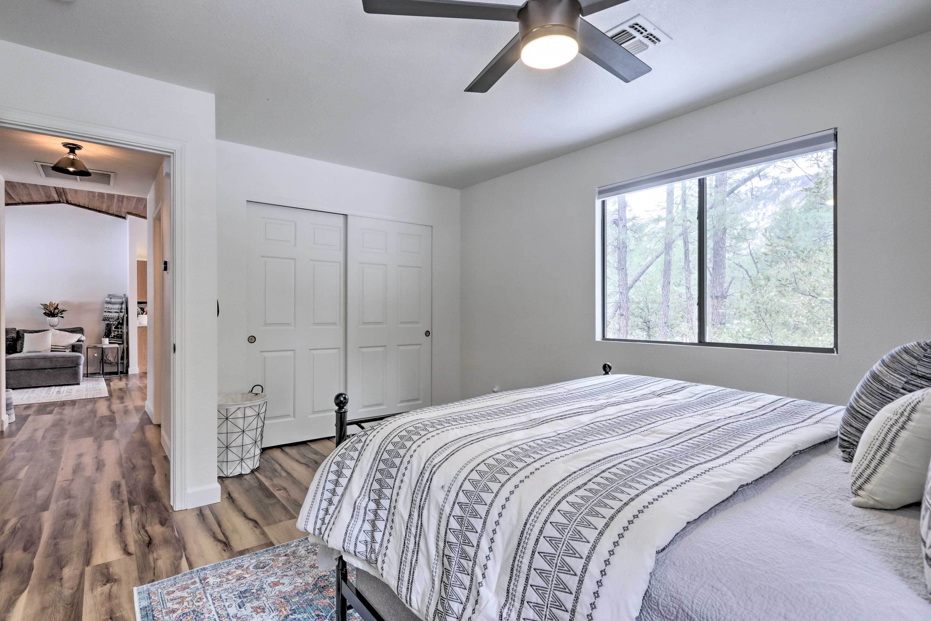 A bedroom with a bed, a ceiling fan, a window with a view of trees, and a door leading to another room.
