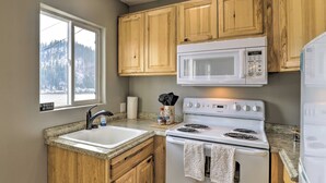 A kitchen with wooden cabinets, a white stove, a microwave, a sink, and a window with a view of trees. - Lake Loon 'hillside' Cottage w/ Private Deck! (Loon Lake)
