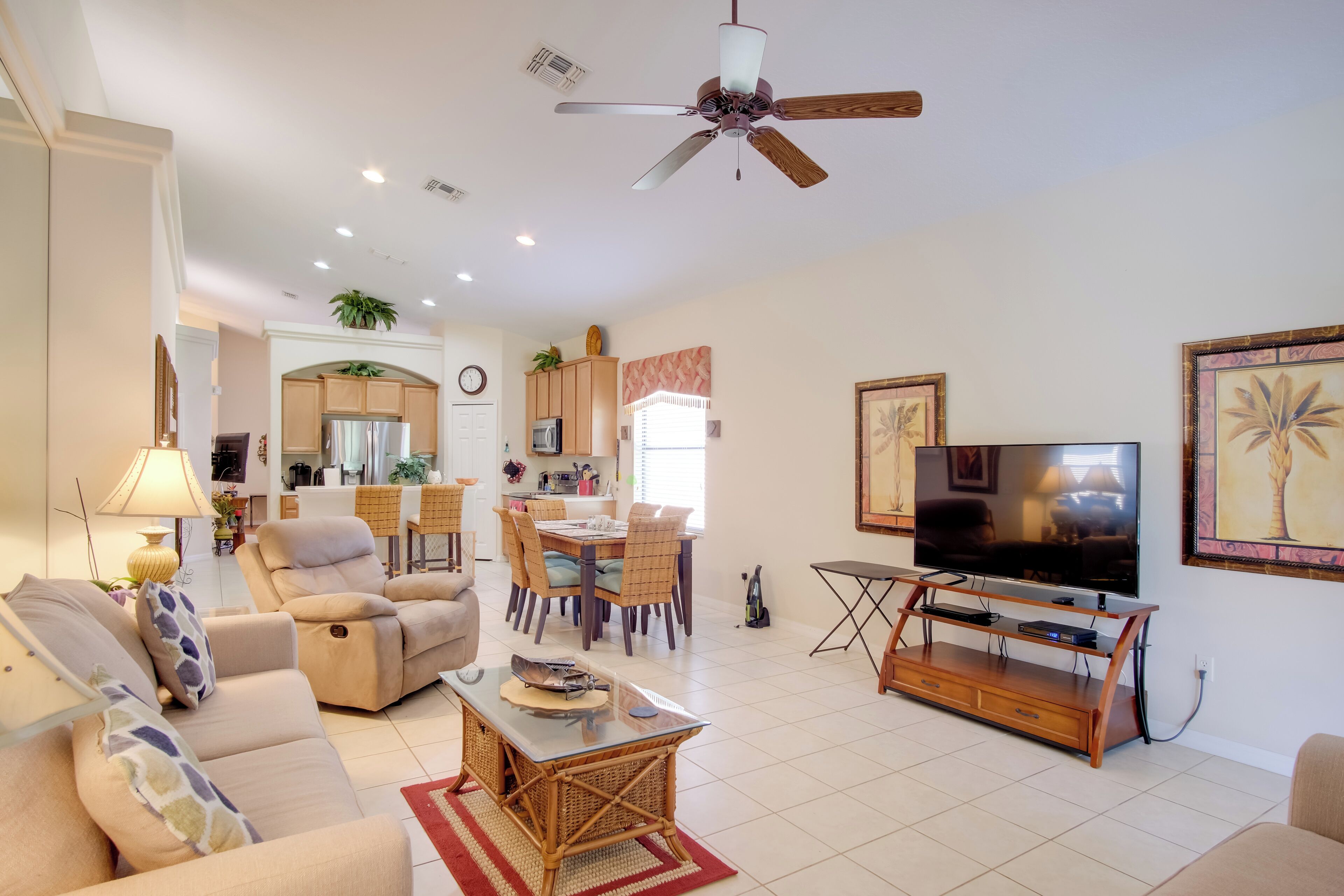A spacious living room with a sofa, armchair, and coffee table, a dining area with chairs and a table, a kitchen with cabinets and appliances, and a ceiling fan.