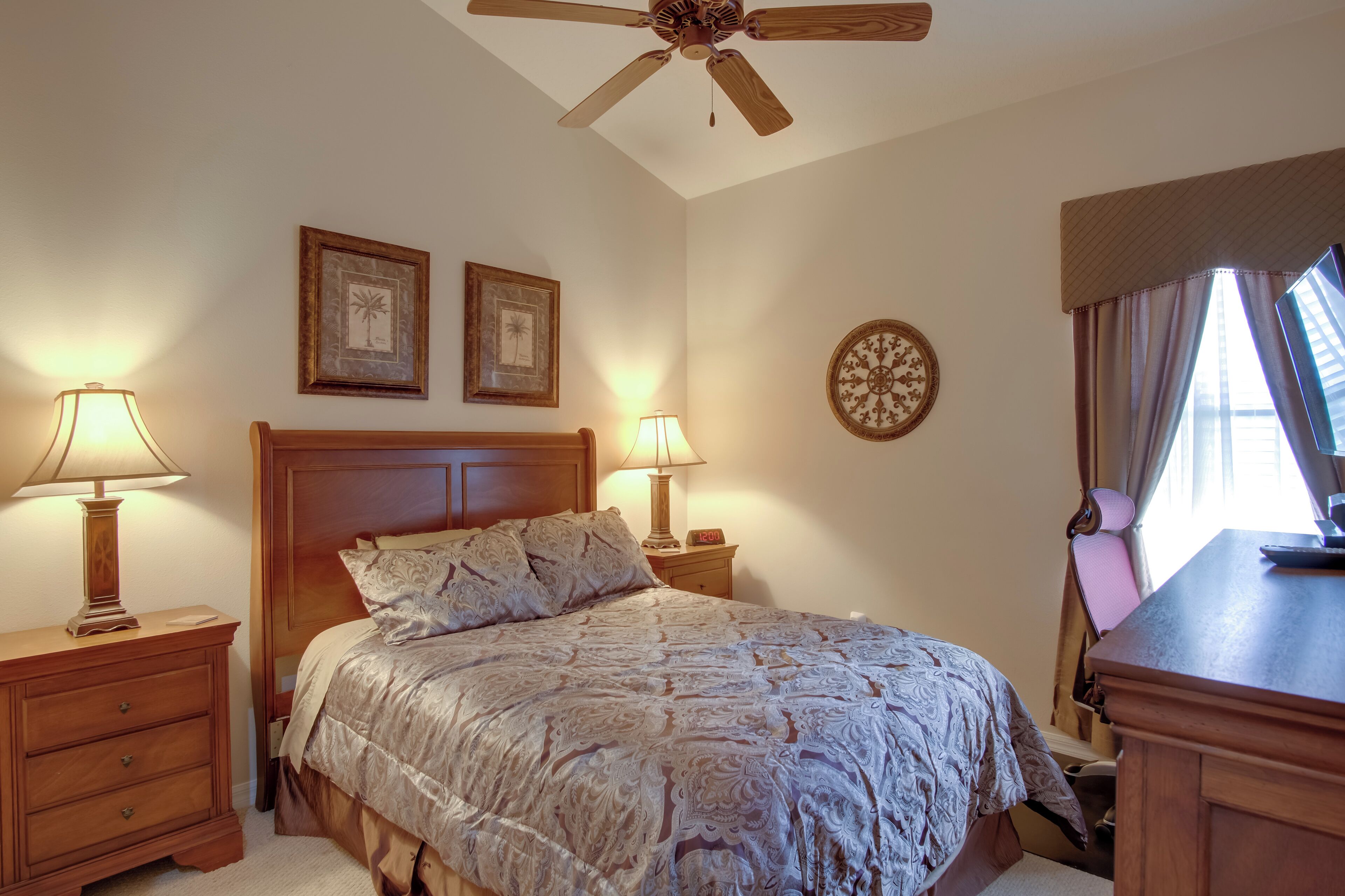 A bedroom with a wooden bed, two bedside tables with lamps, a ceiling fan, and a window with curtains.