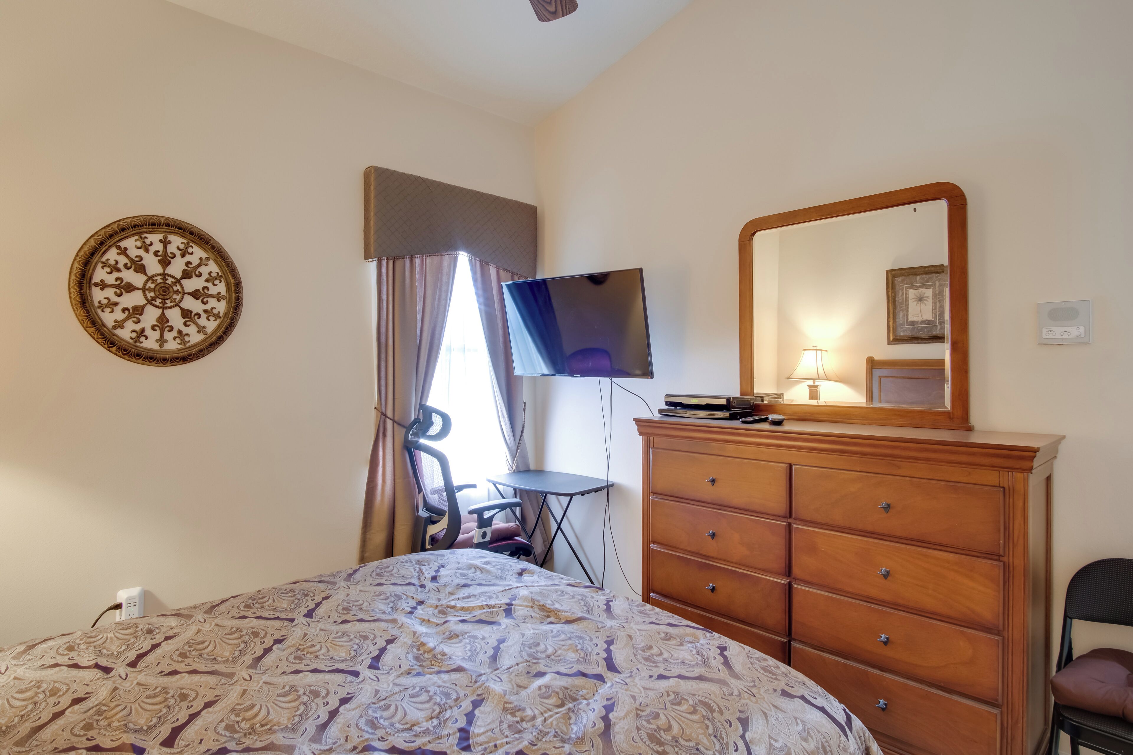 A bedroom with a bed, a dresser, a TV, a mirror, and a decorative wall piece.