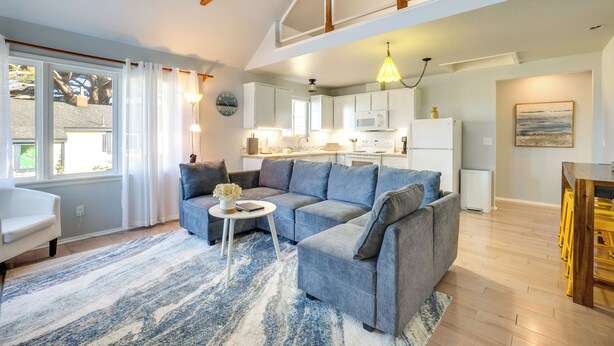 A spacious living area with a grey sectional sofa, a small round coffee table, and a blue and white rug. A kitchen with white cabinets and a microwave is visible in the background. - Bandon Beach House Rental: Walk to Downtown! (Bandon)