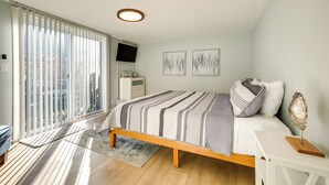 A bedroom with a bed, a bedside table, a TV, and a window with blinds. - Bandon Beach House Rental: Walk to Downtown! (Bandon)