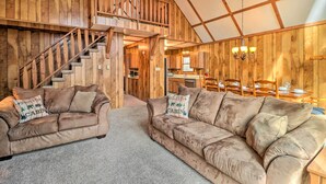 A rustic cabin interior with a wooden staircase, two beige sofas with 'Welcome to Cabin' pillows, a chandelier, and a dining area with wooden furniture. - Lake Harmony Access: Home w/ Game Room + Fire Pit! (Lake Harmony)