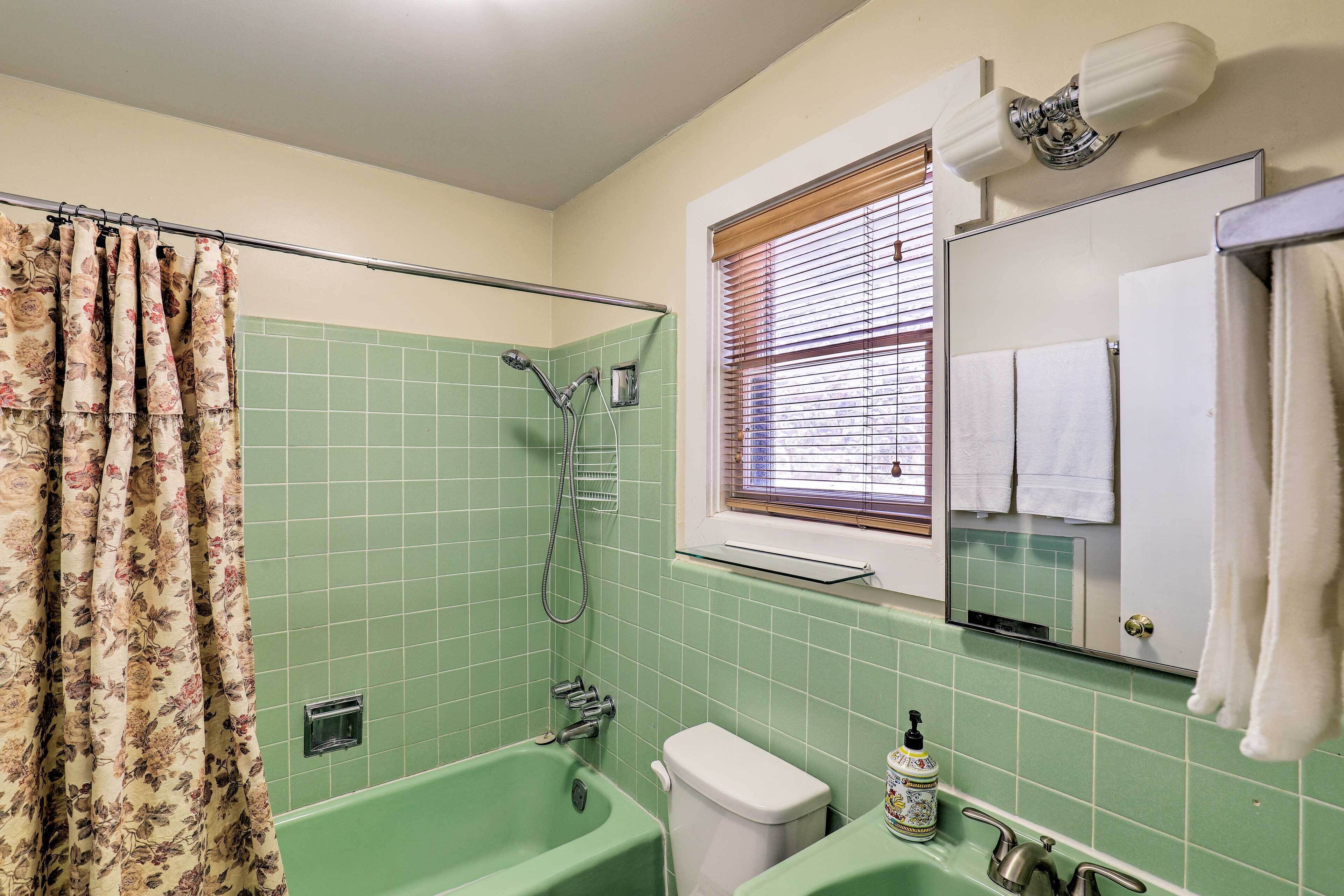 Un baño con azulejos verdes, una bañera, un inodoro, una ducha, una ventana con persianas y un espejo con una toalla colgada.