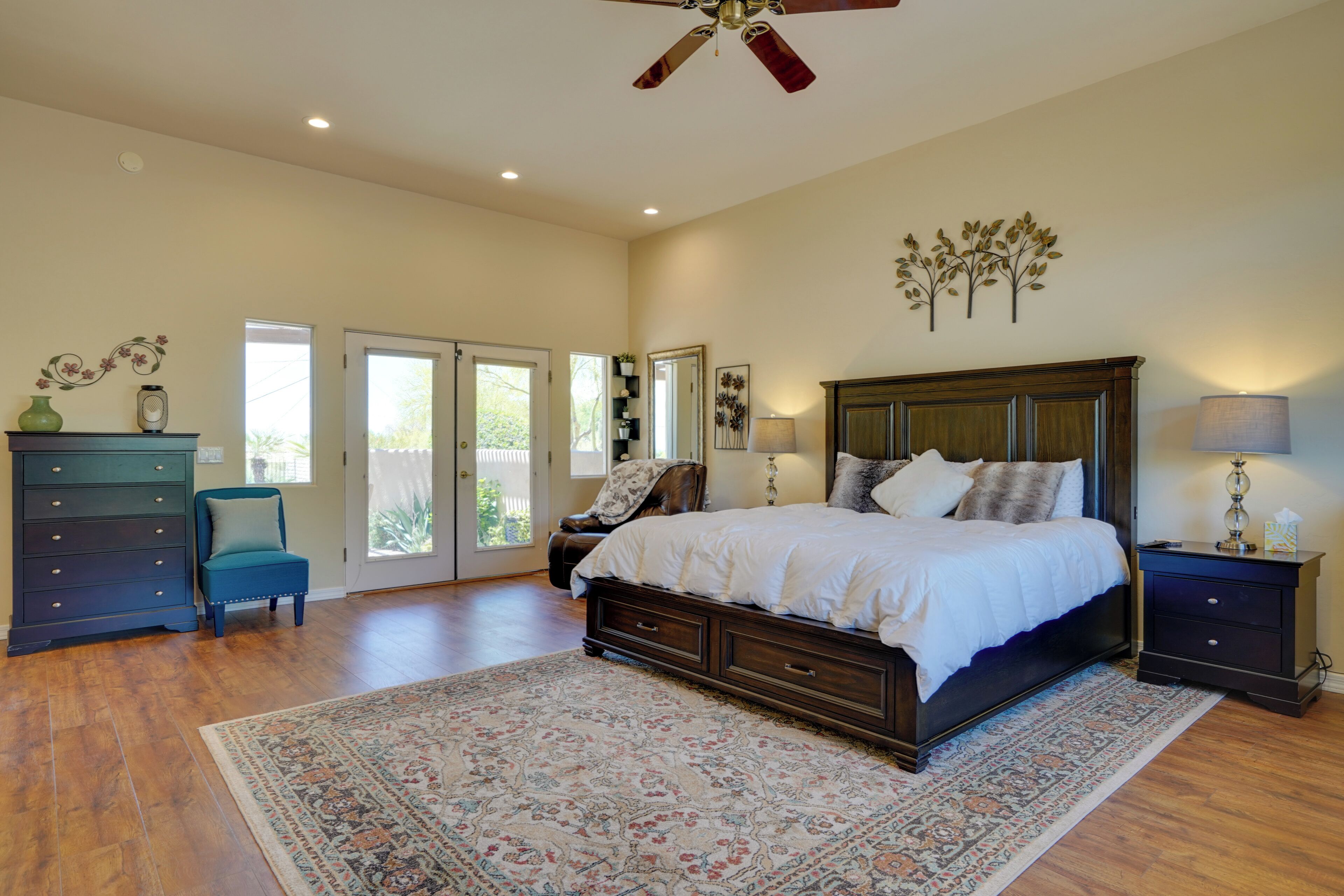 A bedroom with a large bed, a dresser, a chair, a ceiling fan, and a view through a glass door.
