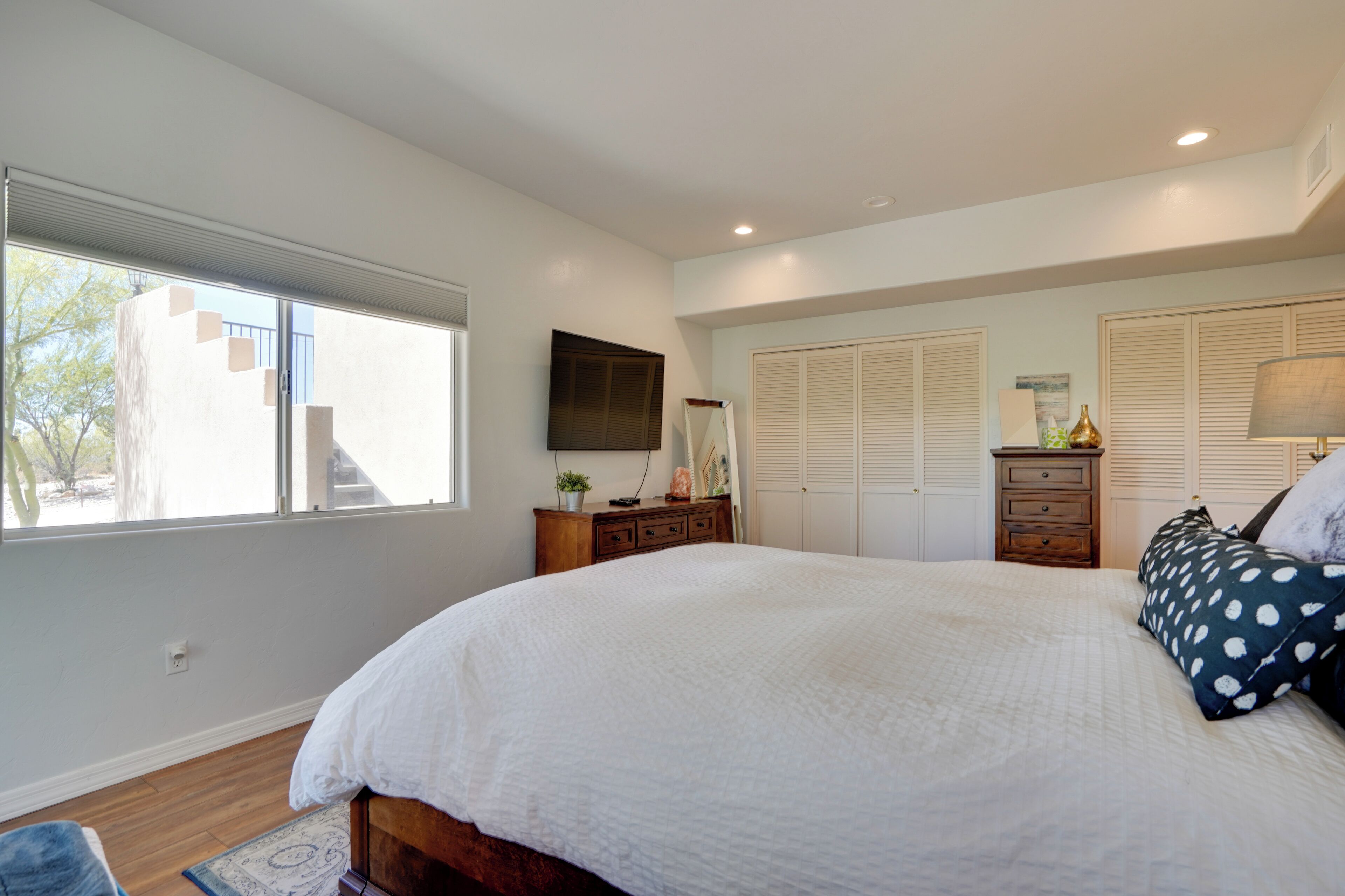 A bedroom with a large bed, a dresser, a nightstand, a lamp, and a window with blinds.