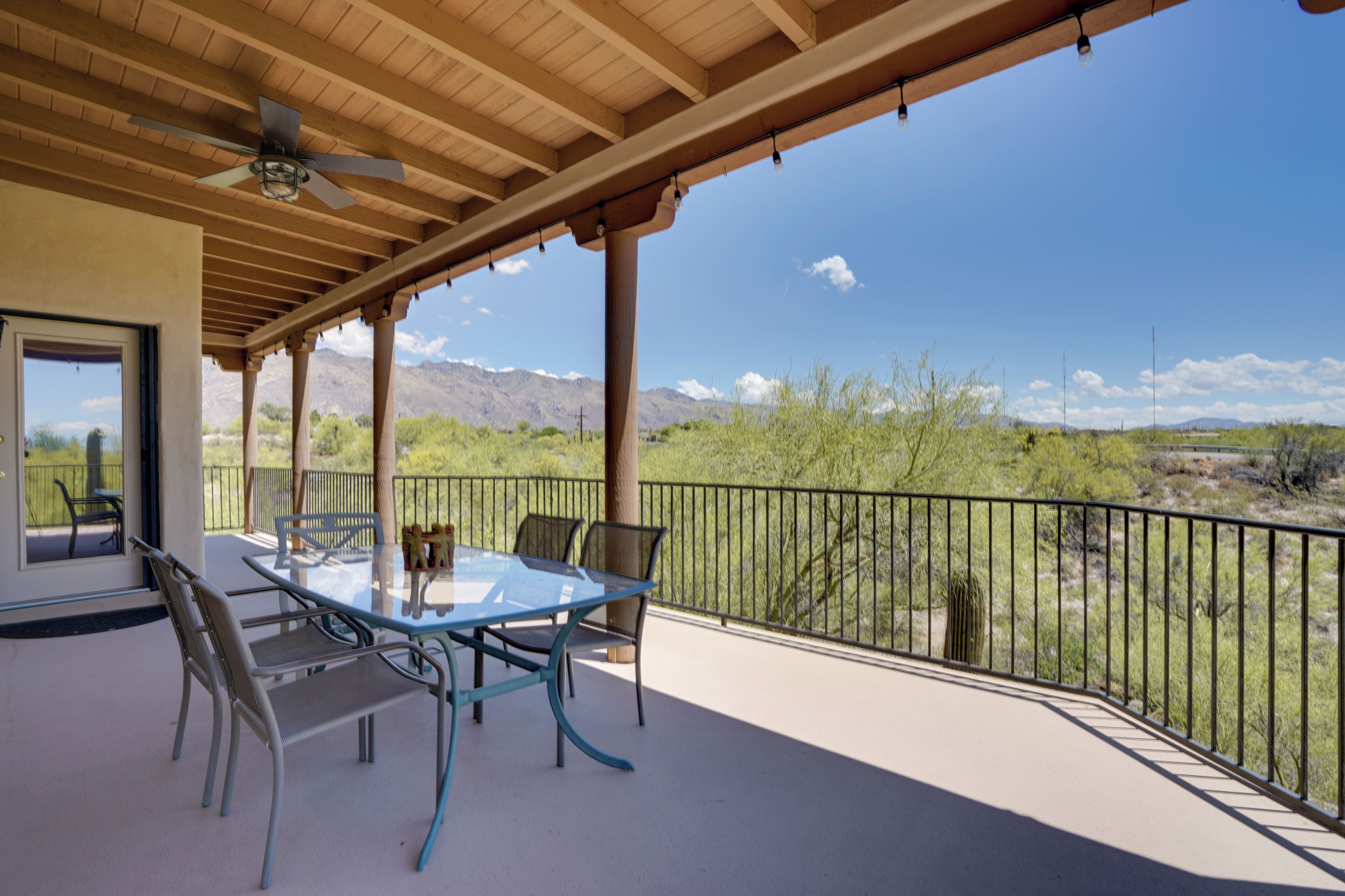 A covered patio with a glass table and chairs, a ceiling fan, and a view of a desert landscape.
