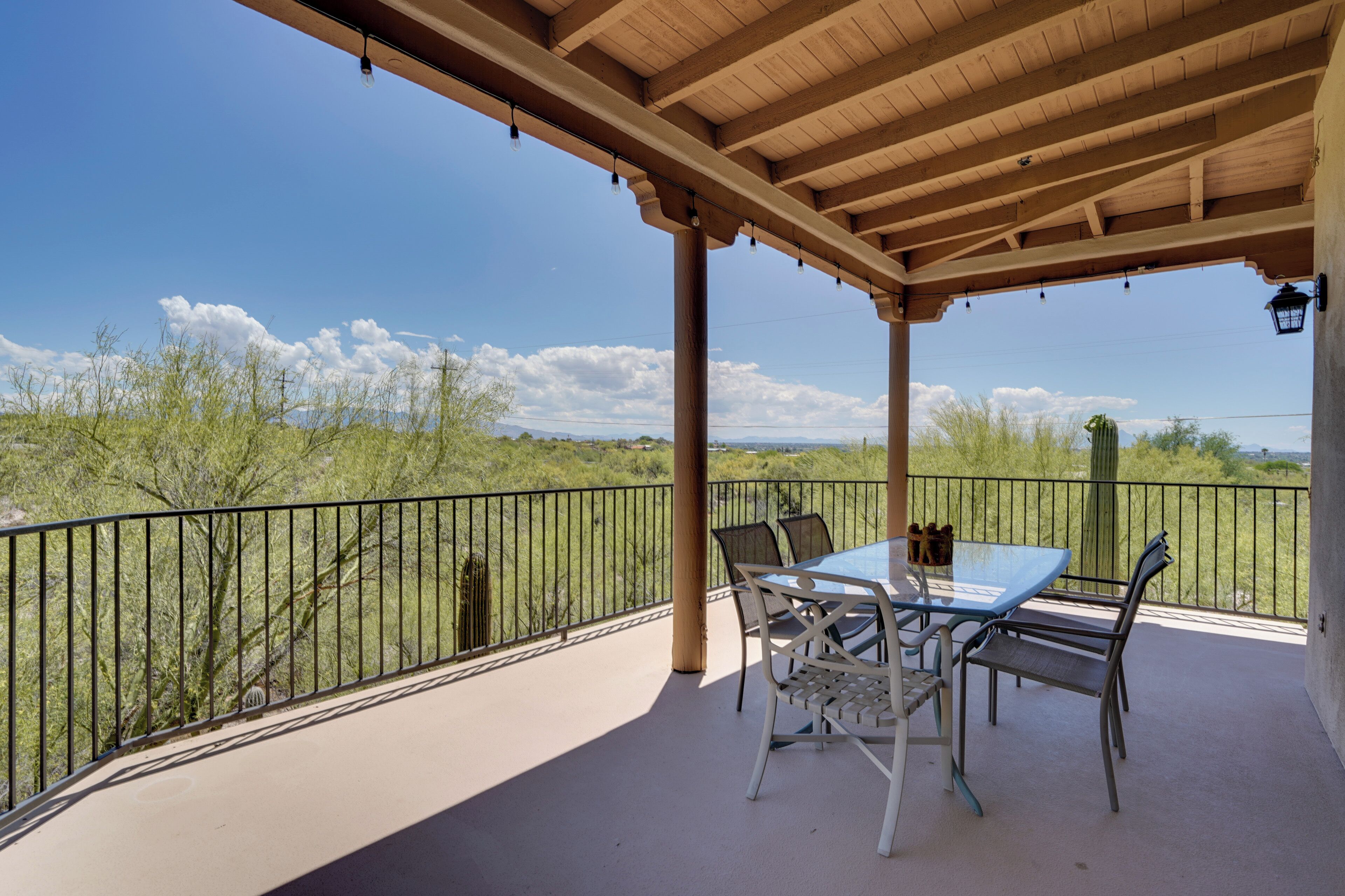 A covered outdoor patio with a table and chairs, a view of the surrounding landscape, and a black metal railing.