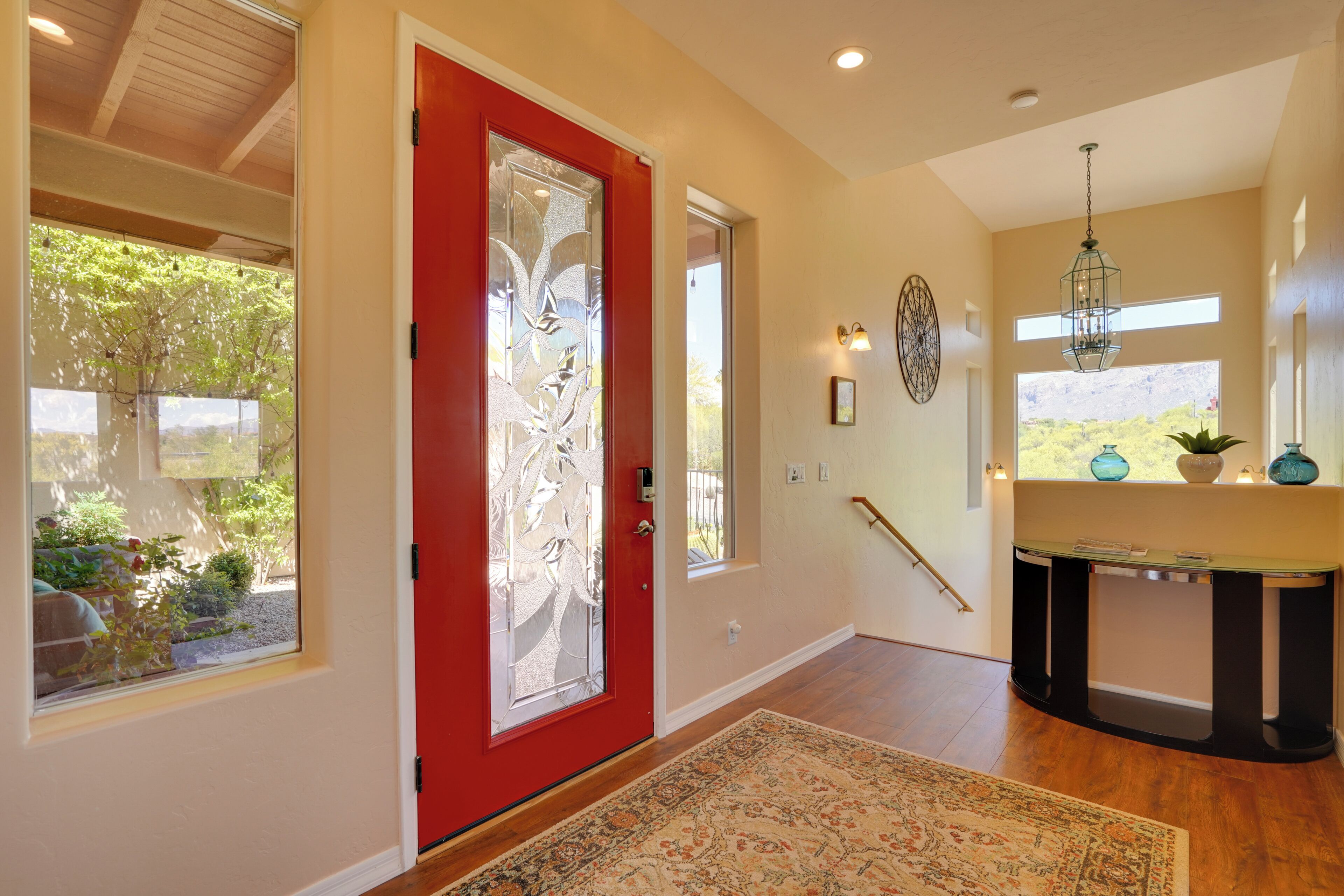 A hallway with a red door, a patterned rug, a console table with decorative items, and a large window with a view of greenery.