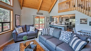 A spacious living area with a high ceiling and wooden beams, a grey sofa set, a coffee table with a plant, and a view of the kitchen and balcony. - Modern Mountain Chalet: Munds Park Retreat w/ Deck (Munds Park)