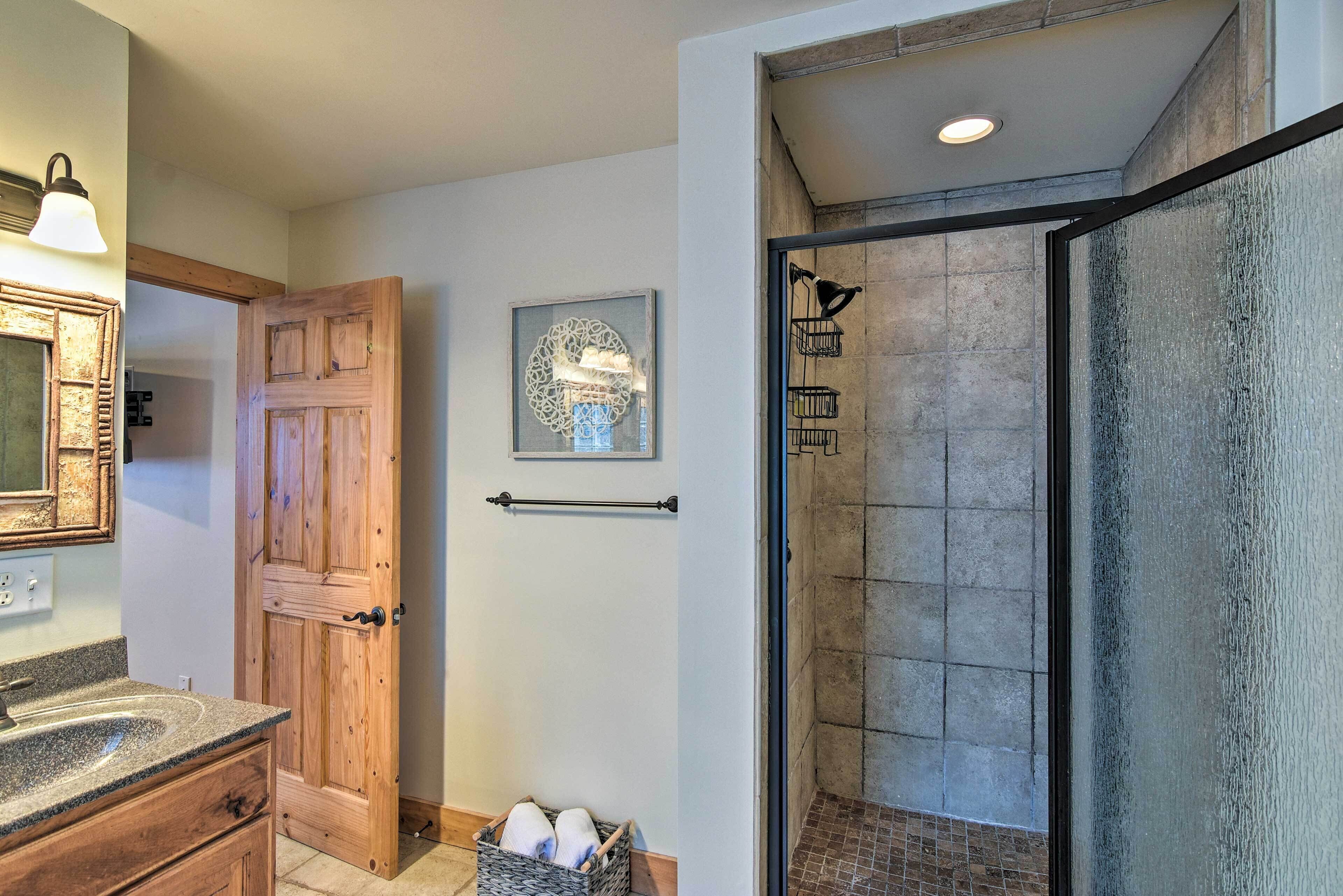 A bathroom with a wooden door, a sink with a granite countertop, a framed artwork above the door, and a shower area with a glass door.