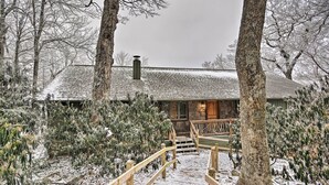 Uma cabana de madeira coberta de neve, com chaminé, cercada por árvores e um deck de madeira.