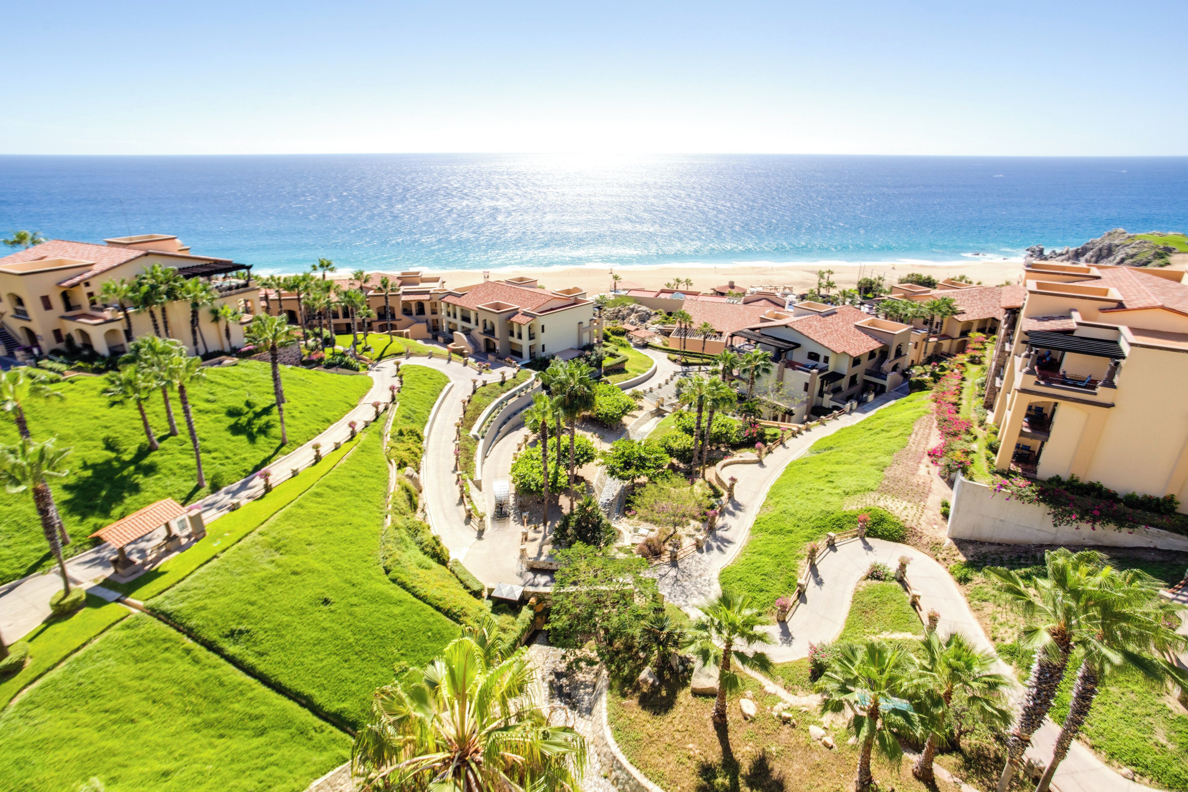 A resort with a beachfront view, featuring a central courtyard with palm trees and pathways, and buildings with red-tiled roofs.