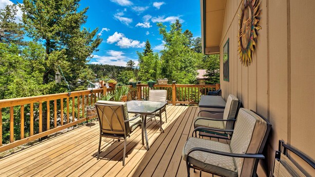 Una terraza de madera con mobiliario exterior, barandilla y vistas a árboles y montañas.