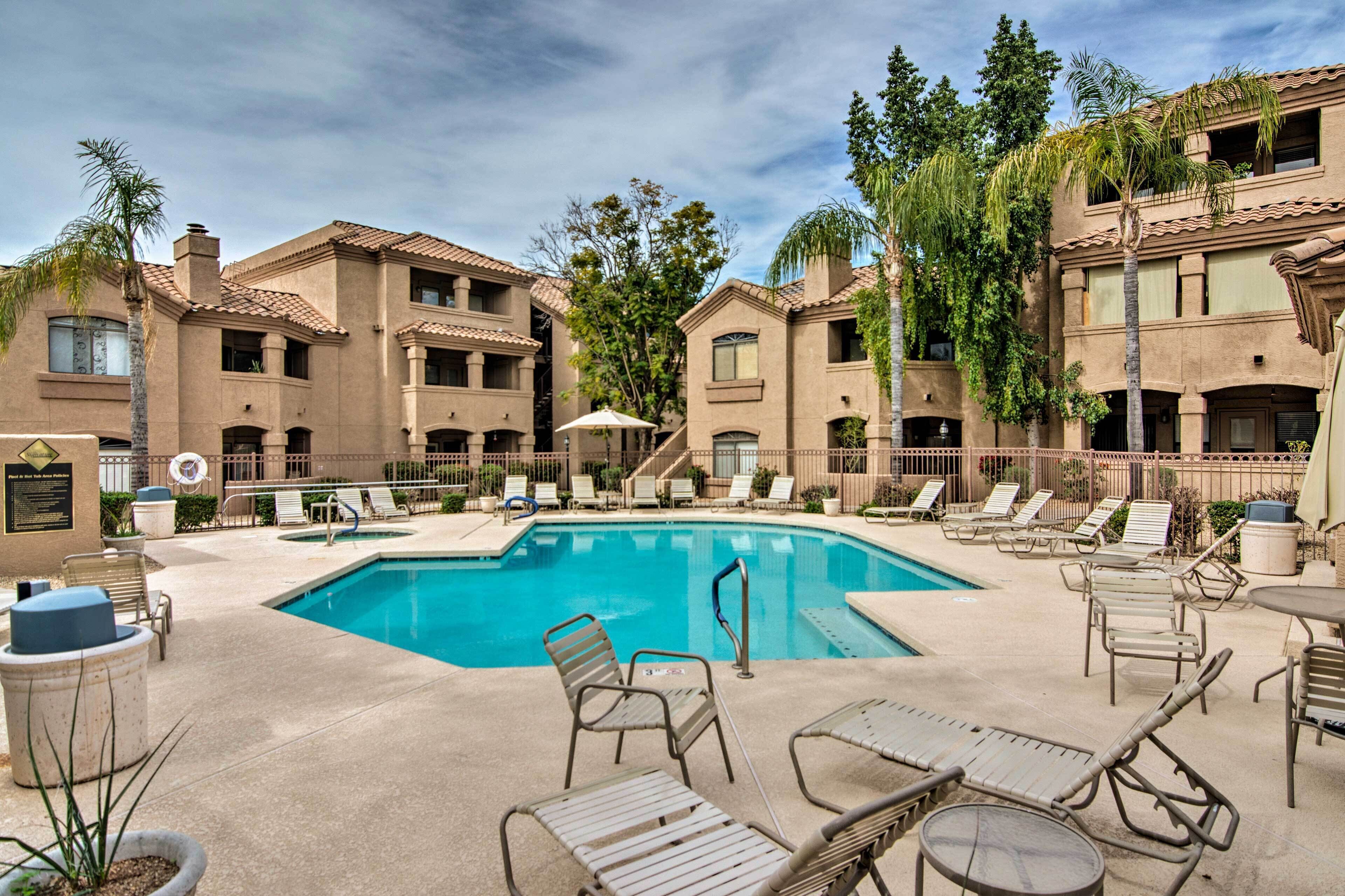 A pool area with lounge chairs and a clear blue pool surrounded by buildings with balconies and palm trees.