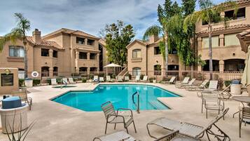A pool area with lounge chairs and a clear blue pool surrounded by buildings with balconies and palm trees.