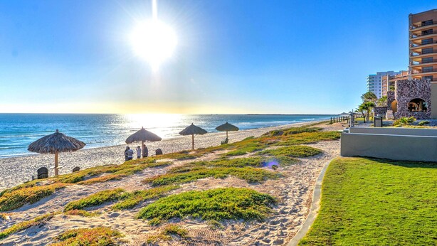 A beach with umbrellas, a building with a stone facade, and a clear sky. - Beachfront Rocky Point Villa w/ Ocean Views! (Puerto Peñasco)