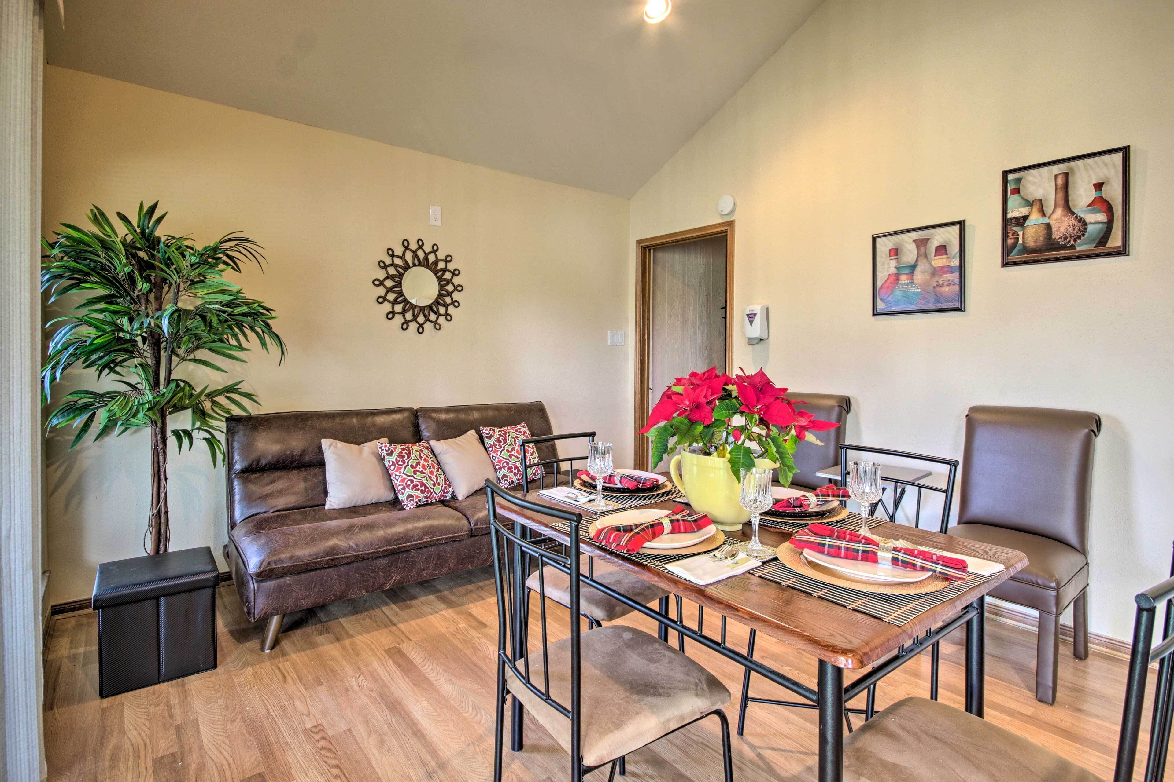 A dining area with a wooden table set for four, surrounded by chairs, a brown leather sofa, and a potted plant.