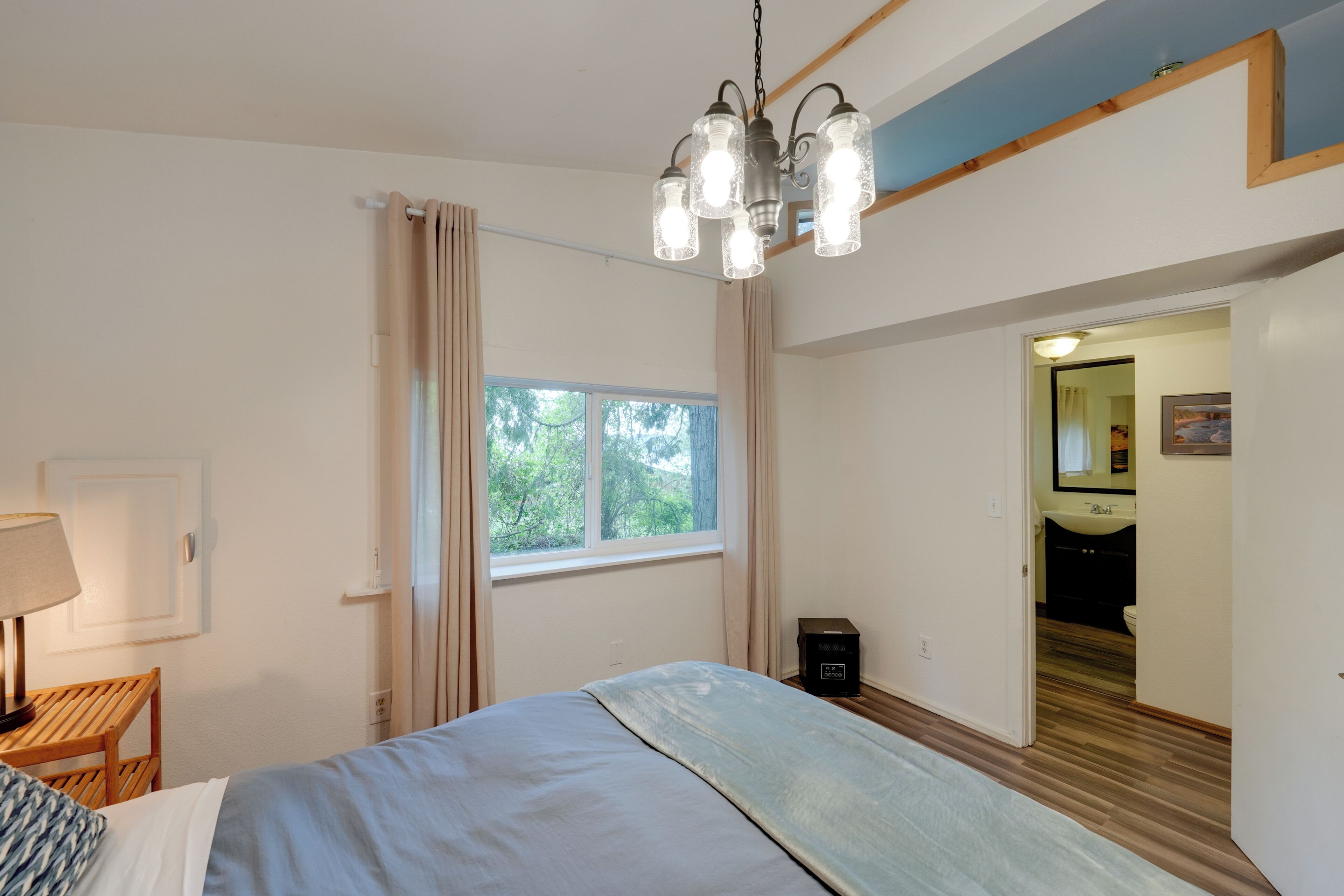 A bedroom with a bed, a bedside table, a lamp, a window with curtains, and a chandelier.