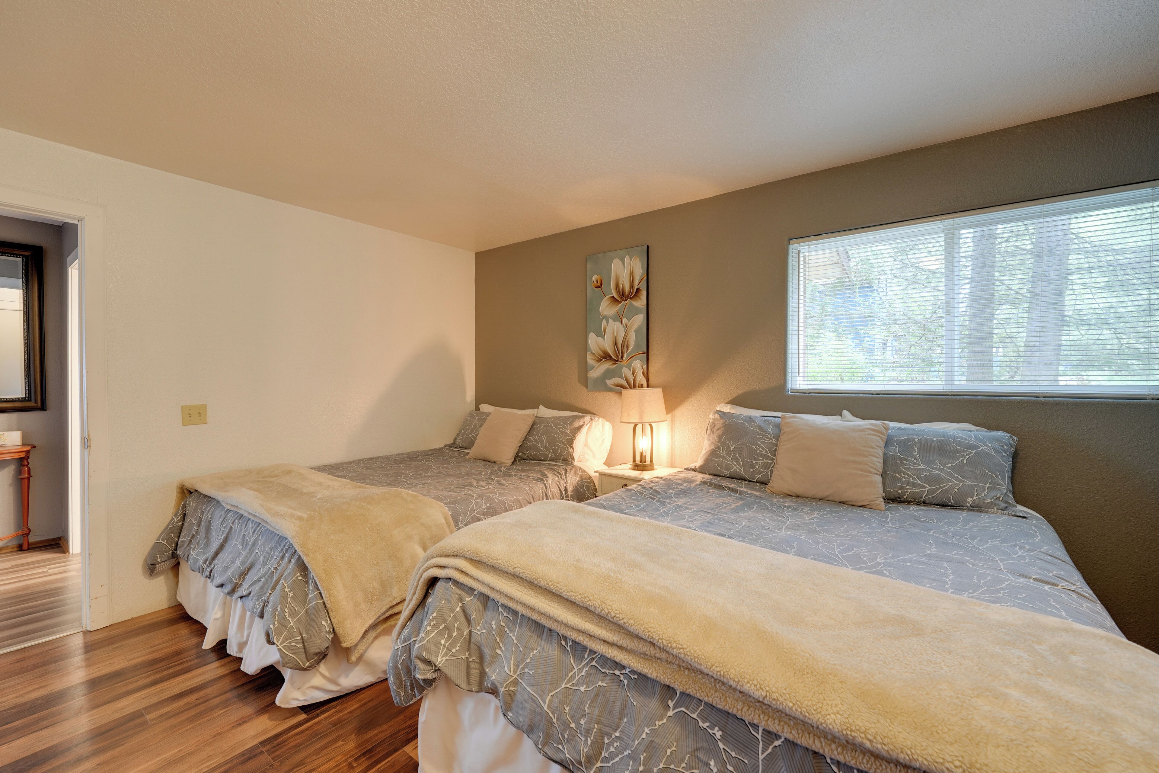 A bedroom with two beds, a nightstand with a lamp, a painting on the wall, and a window with blinds.