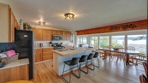 A spacious kitchen with wooden cabinets, a central island with bar stools, a dining area with wooden chairs, and large windows offering a view. - Cozy Beachouse: View & Deck, Steps From Skagit Bay (La Conner)