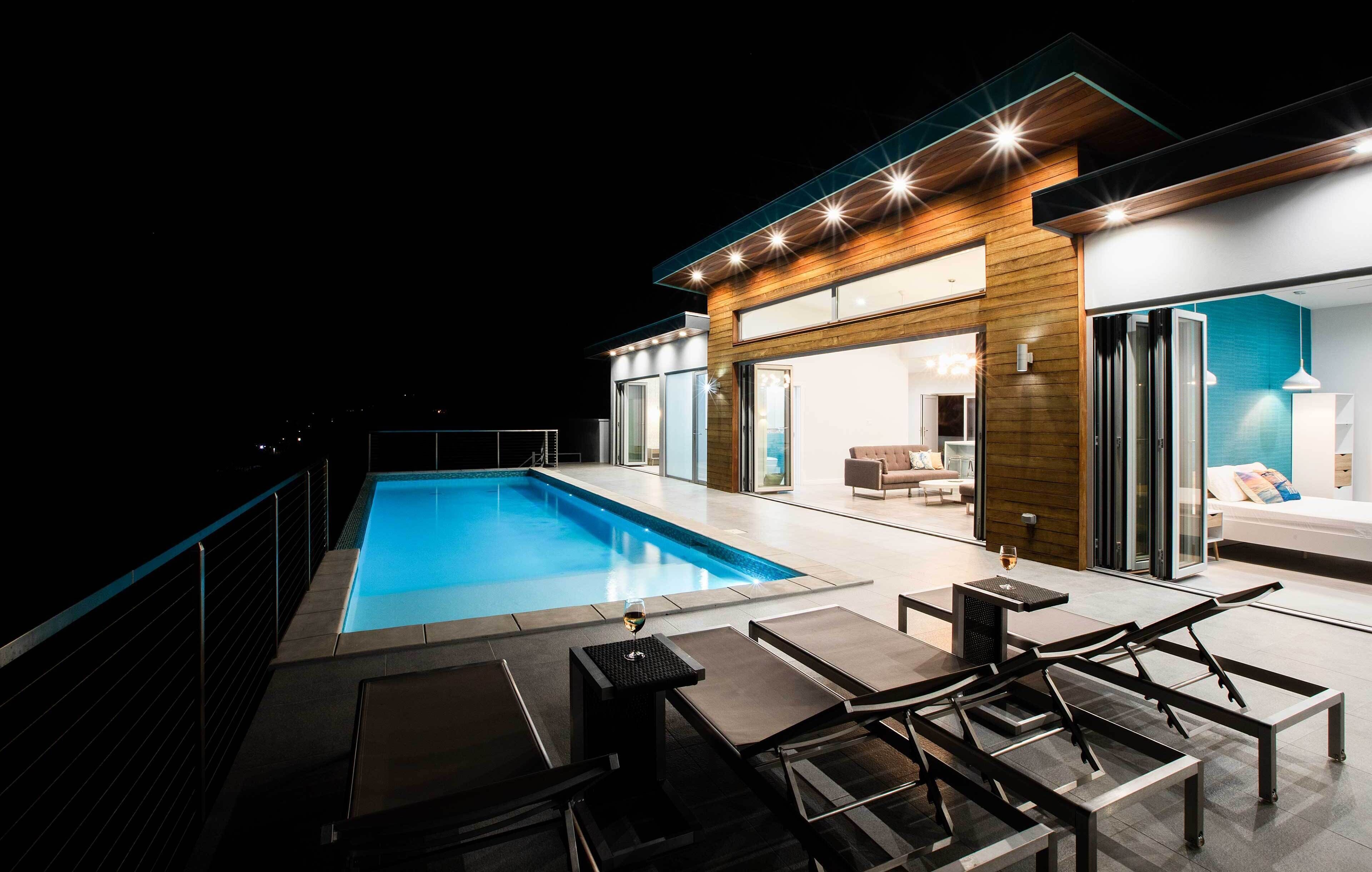 A modern outdoor pool area with a glass enclosure, lounge chairs, and a table with a glass of wine.