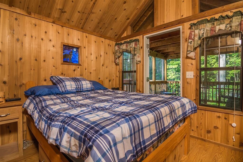A wooden cabin bedroom with a plaid bedspread, a nightstand, a small table, and a window with a view of greenery.