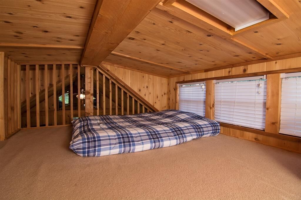 A wooden room with a bed, windows with blinds, and a sloped ceiling.