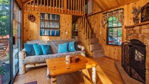 A cozy cabin interior with a wooden ceiling, a sofa with patterned cushions, a coffee table, a fireplace, and a large window offering a view of the outdoors. - Family Cabin on 6 Acres w/ Lake Access & Hot Tub! (Springfield)