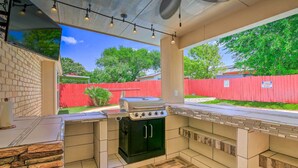 Une cuisine extérieure équipée d’une cuisinière à gaz, d’un barbecue et d’un four encastré, le tout entouré d’une clôture rouge et d’arbres.