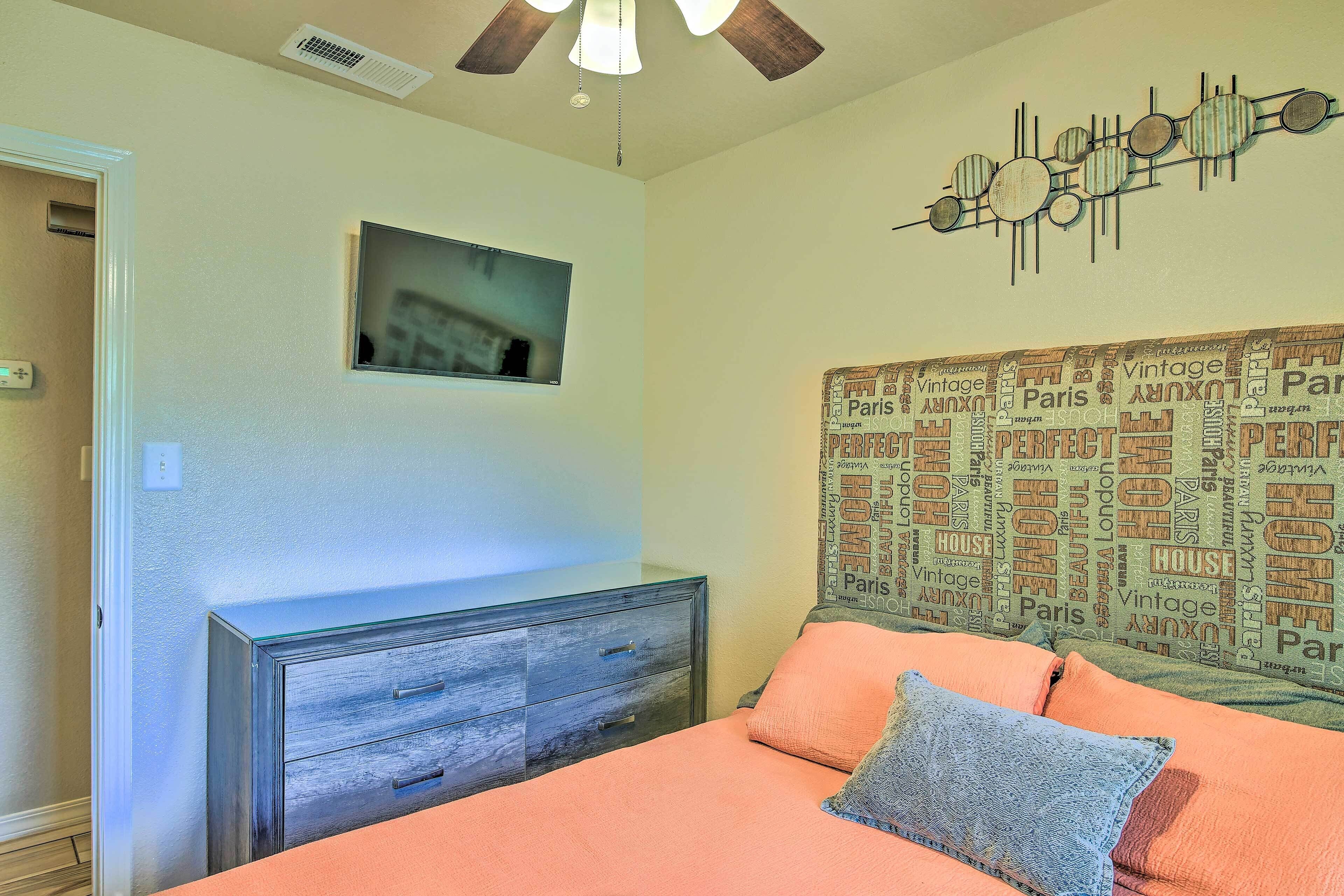 A bedroom with a bed, a dresser, a ceiling fan, a wall-mounted TV, and a decorative wall piece.