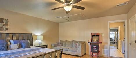 A bedroom with a bed, a ceiling fan, a nightstand, a lamp, a couch, and a small arcade machine.