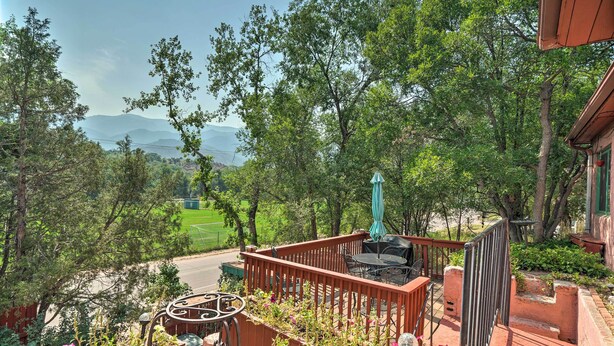 A wooden deck with a table and chairs, surrounded by greenery and a view of distant mountains. - Colorful Bungalow By Pikes Peak/garden of the Gods (Manitou Springs)