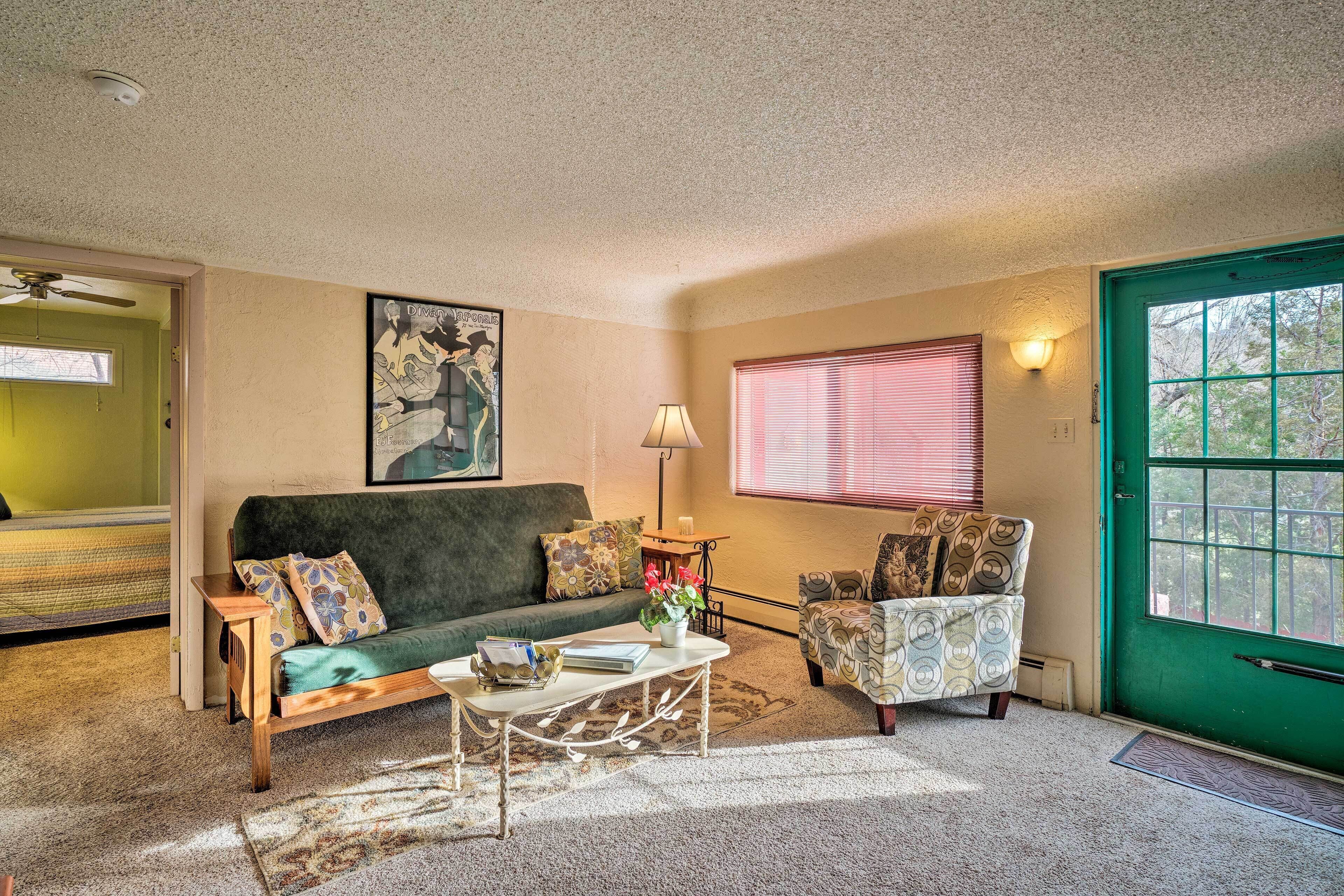Una sala de estar acogedora con una puerta verde, un sofá, un sillón estampado, una mesa de centro con libros y un jarrón de flores, y un cuadro en la pared.